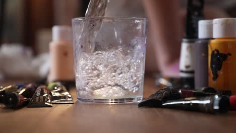 slow-motion of water poured in glass for painting purposes. camera moves away from the glass as the water is being poured, painting tubes and instruments.