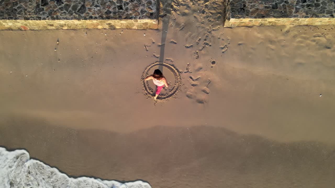 태국에서 그녀의 뒤에 부서지는 파도와 함께 모래에서 원을 만드는 한 여성의 해변을 직접 내려다보는 공중 전망