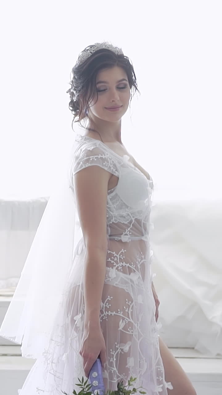 Donna meravigliosa in biancheria intima di pizzo con una tiara brillante in piedi davanti a un lungo abito da sposa bianco al rallentatore.