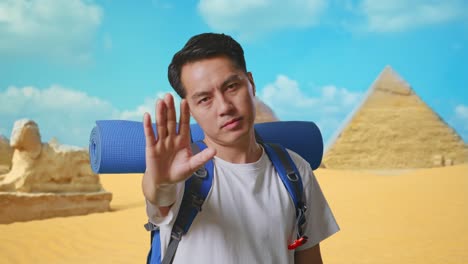 close up of asian male hiker with mountaineering backpack looking at camera and making stop hand sign while traveling in pyramid of giza