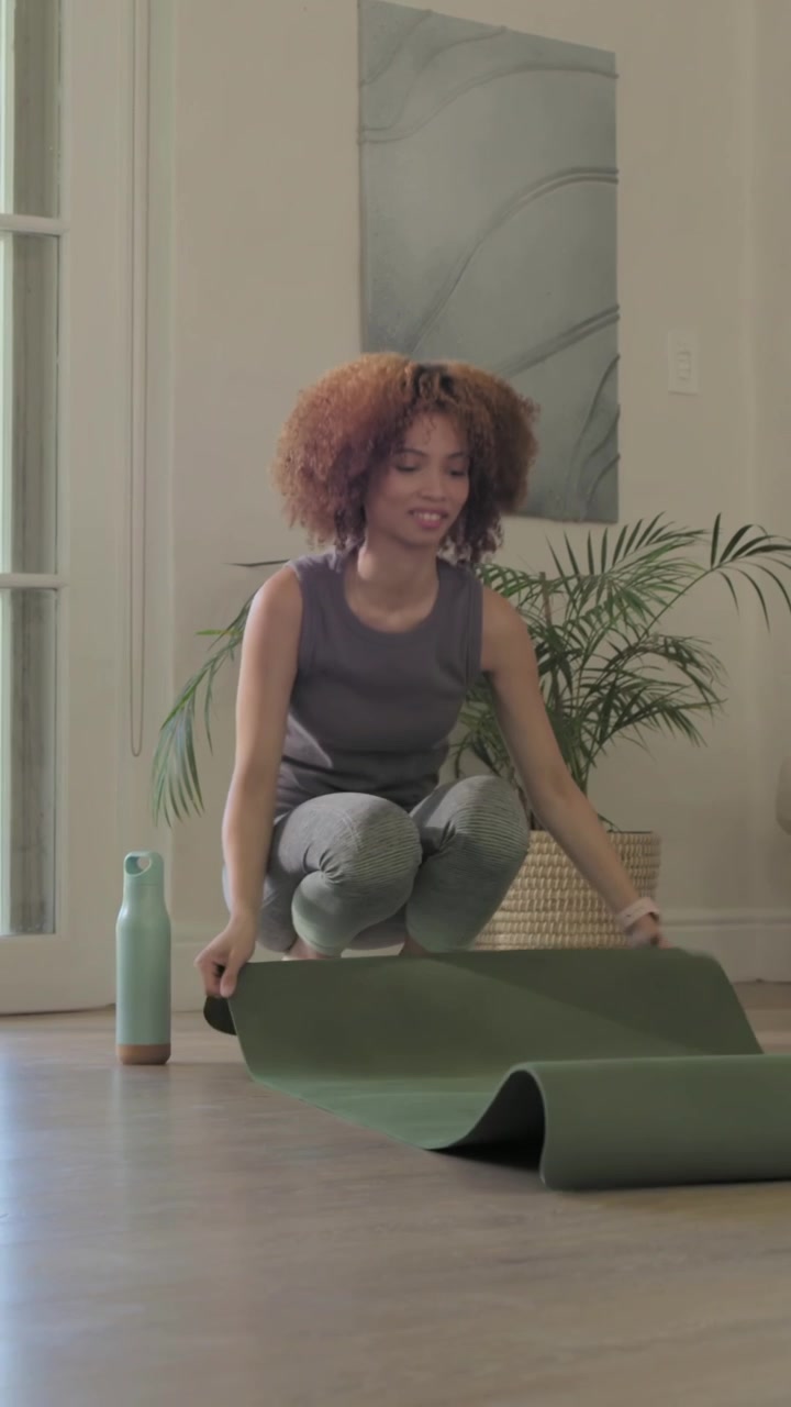 Video verticale: Arrivando con i tappetini, donna che srotola il tappetino a casa in leggings e canotta, preparandosi per lo yoga.