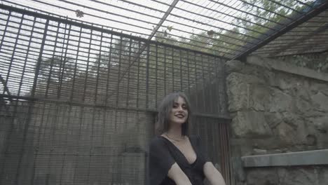 woman with expressive make-up and black dress stares amusedly at the sky through the bars of the cage in which she is placed