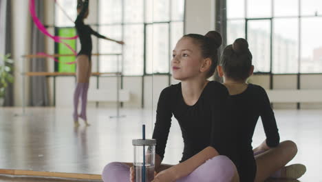 gymnastic blonde girl sitting on the floor while watching to her friend dancing holding a band