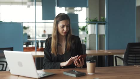 una mujer joven y bonita con ropa de negocios se sienta en una elegante oficina moderna de coworking, molesta por los mensajes en su teléfono inteligente que la distraen del trabajo en su portátil