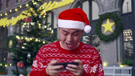 close up of asian man in santa suit celebrating winning game on smartphone while standing in christmas night