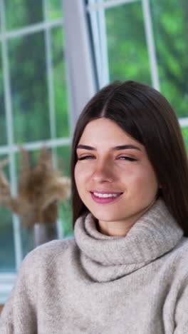 attractive brunette in warm sweater indoors. young lady touching her hair, posing and smiling for camera. modern clothes and footwear presentation. vertical video