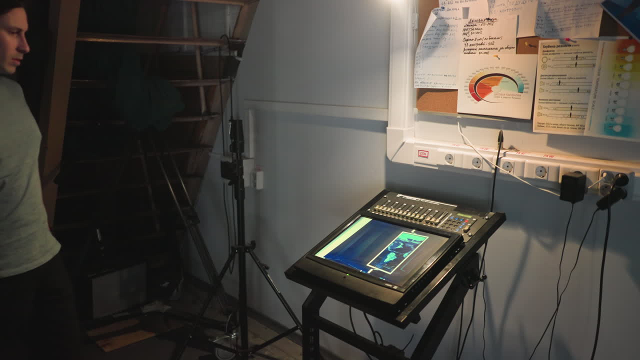 Young male technician operates digital control console in dimly lit backstage room. Screen displays interface graphics under focused desk lamp. Surrounding notes and outlets create technical, professional mood