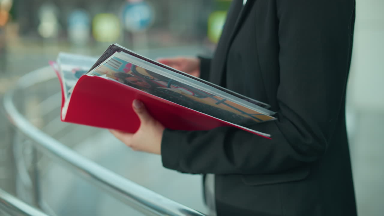 Side view of woman in black blazer browsing through her colorful artistry book, holding red folder with creative works, with soft blur background of urban setting and metal railing in natural light