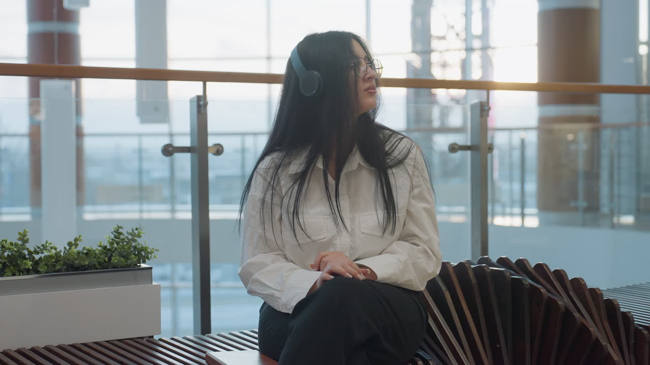 Exhausted woman with headphones and glasses sitting on modern wooden bench in mall interior shuts hardback book, sets it aside, crosses legs, lifts head toward windows, seeking inspiration