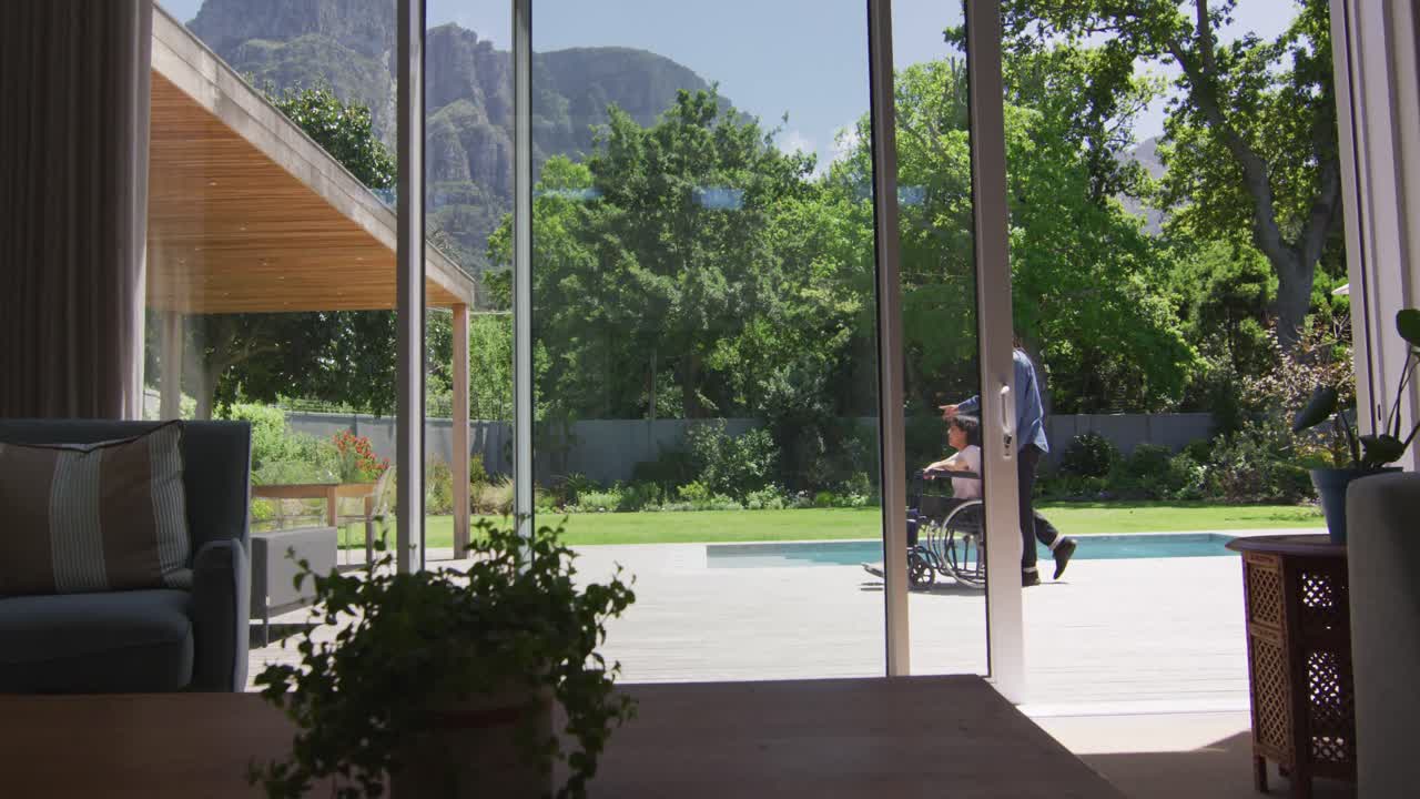 hombre biracial con dreadlocks empujando a su pareja femenina en silla de ruedas en un jardín soleado