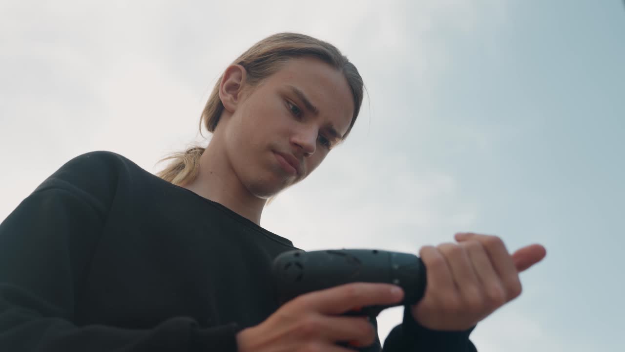 Jeune homme préparant une perceuse électrique en extérieur sous un ciel ouvert, un créateur urbain teste la batterie et le couple, ambiance d'entrepreneur indépendant, gros plan sur les mains ajustant le foret, expression confiante, composition de vie moderne.