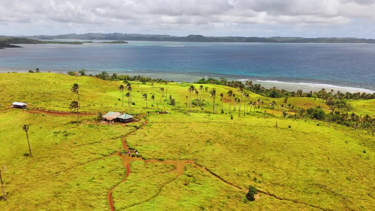 Aerial view of Corregidor Island, Siargao Islands, Philippines shows grassy terrain, palm trees, coastal huts, Pacific Ocean, and surrounding islets under bright daylight in tropical conditions