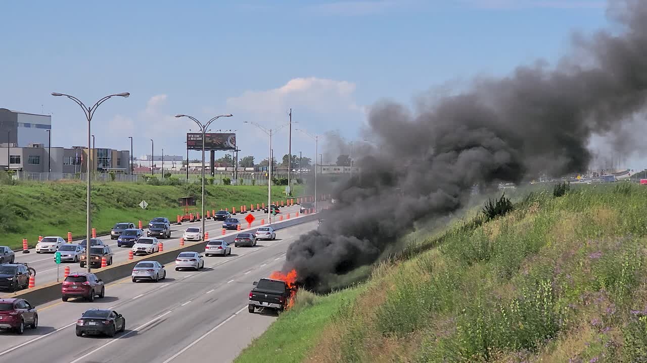 A black SUV on fire by a busy Montreal highway, smoke rising, with passing cars