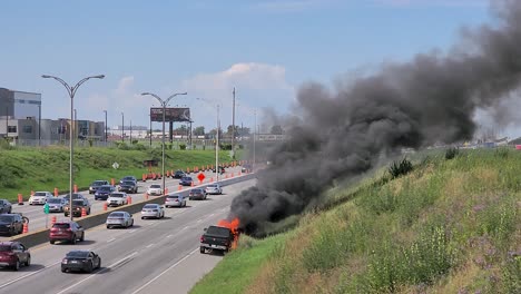 a black suv on fire by a busy montreal highway, smoke rising, with passing cars