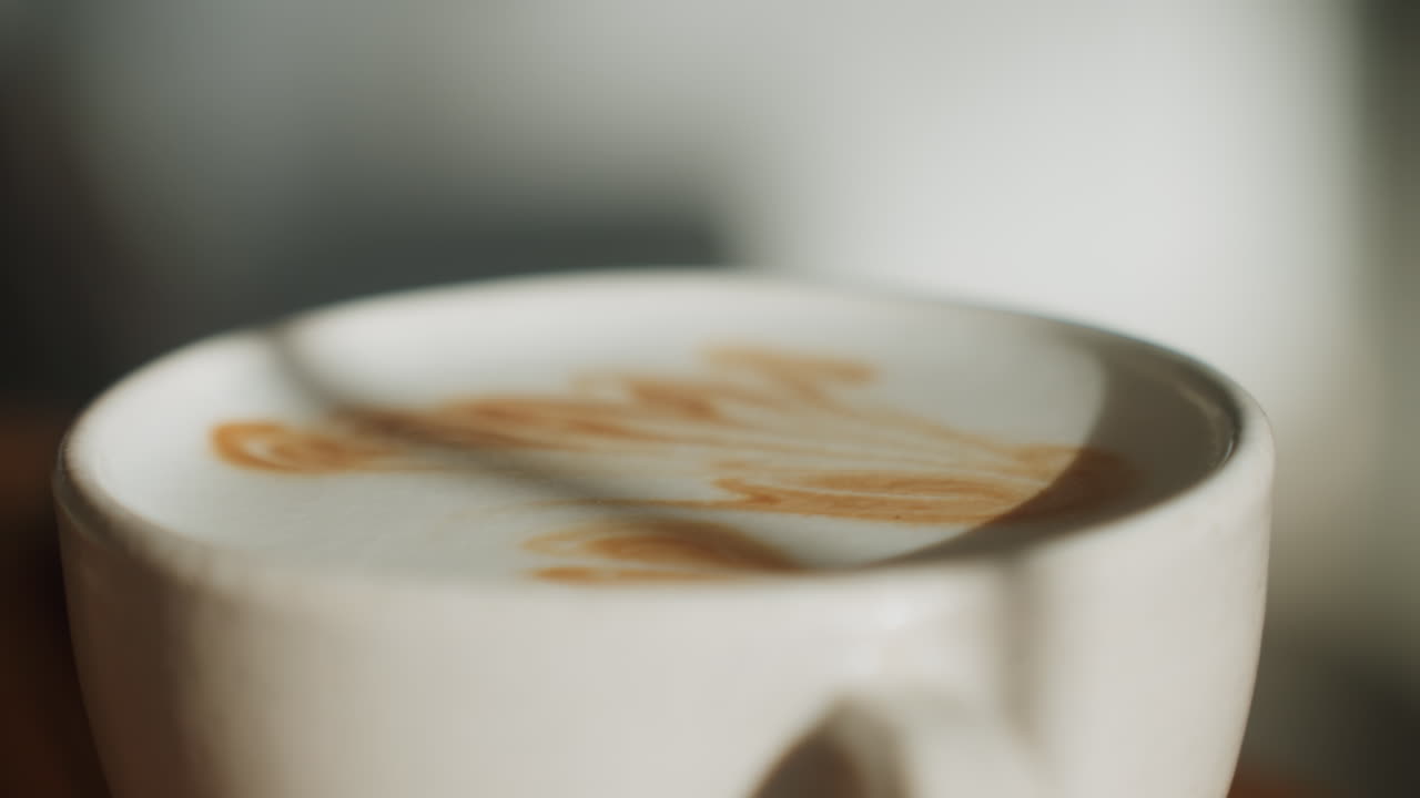 Primo piano cinematografico di schiuma di latte con bokeh drammatico, motivo di crema vellutata che si avvolge sotto un'ombra soffusa, profondità di campo ridotta e sfondo sfocato del caffè, toni caldi e grana cinematografica, caffè intimo.
