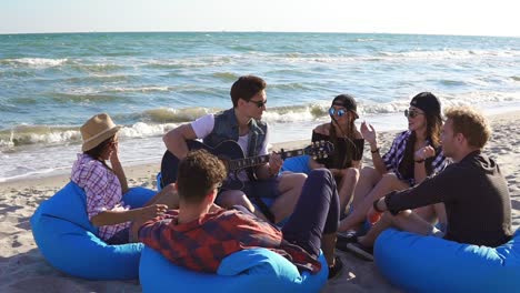 Young-man-playing-guitar-among-group-of-friends-sitting-on-easychairs-on-the-beach-and-singing-on-a-summer-evening.-Slowmotion-shot