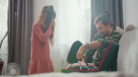 a little brunette girl in a pink dress and her father, a brunette man in a green checkered shirt, put their things in a black suitcase and close it before starting their trip