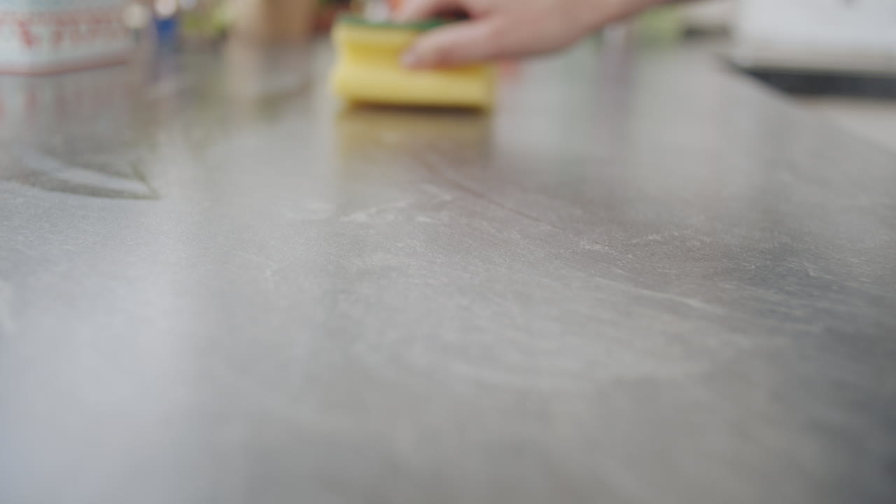 Close up shot of cleaning the kitchen counter with a yellow sponge. caucasian hands wiping from left to right. Camera dollies back. Slow motion shot with shallow depth of field