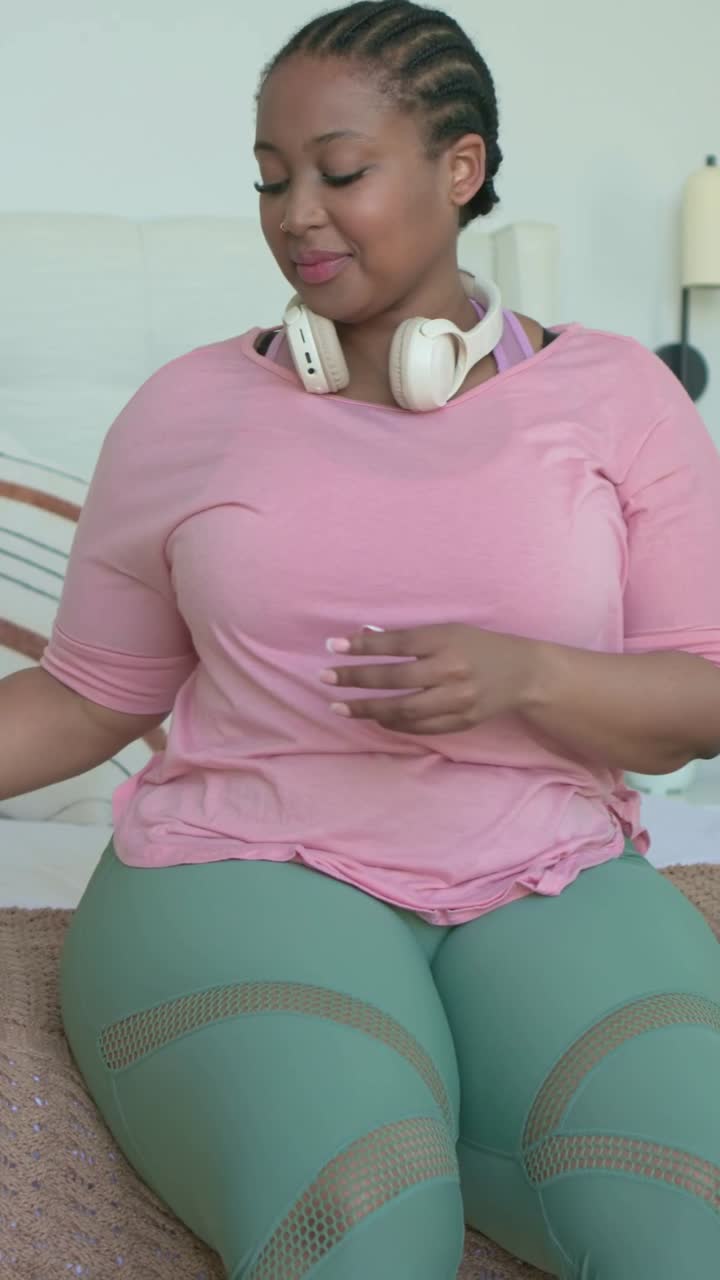 Video verticale: Donna con top rosa che indossa le cuffie, controlla il telefono e beve da una bottiglia rosa sul letto.