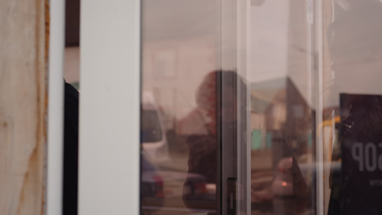 Young man with short hair wearing black jacket and backpack walking into a wooden building, visible neck tattoo, glasses, winter environment, urban background, another man walks with him, casual lifestyle