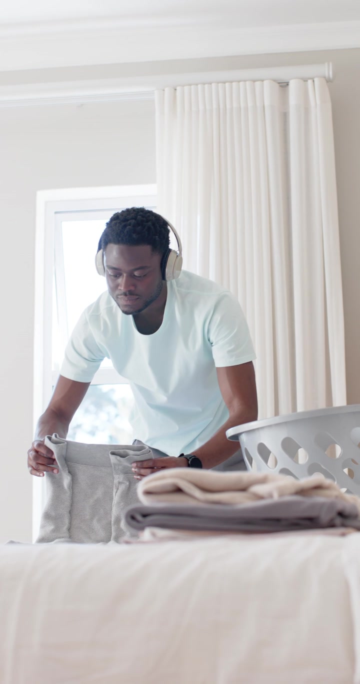 Vertical video: African American man entering bedroom, folding clothes from basket on bed to tidy