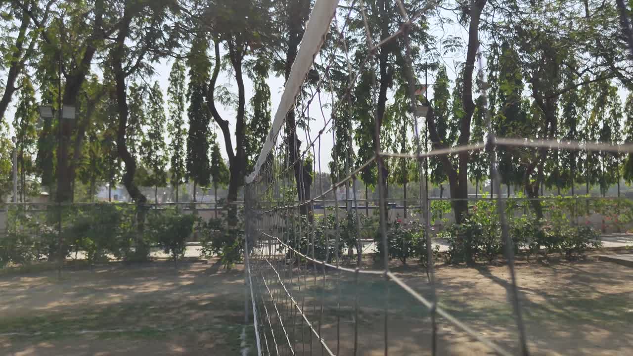 View of volleyball net on an outdoor court, surrounded by lush green trees and sunlight.