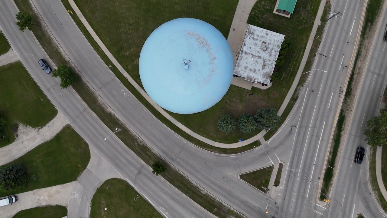 Dron mirando directamente hacia abajo sobre la torre de agua azul claro en Steinbach, Manitoba, avanzando y ascendiendo para revelar la torre del embalse, carreteras, coches, casas, árboles y césped verde en verano.