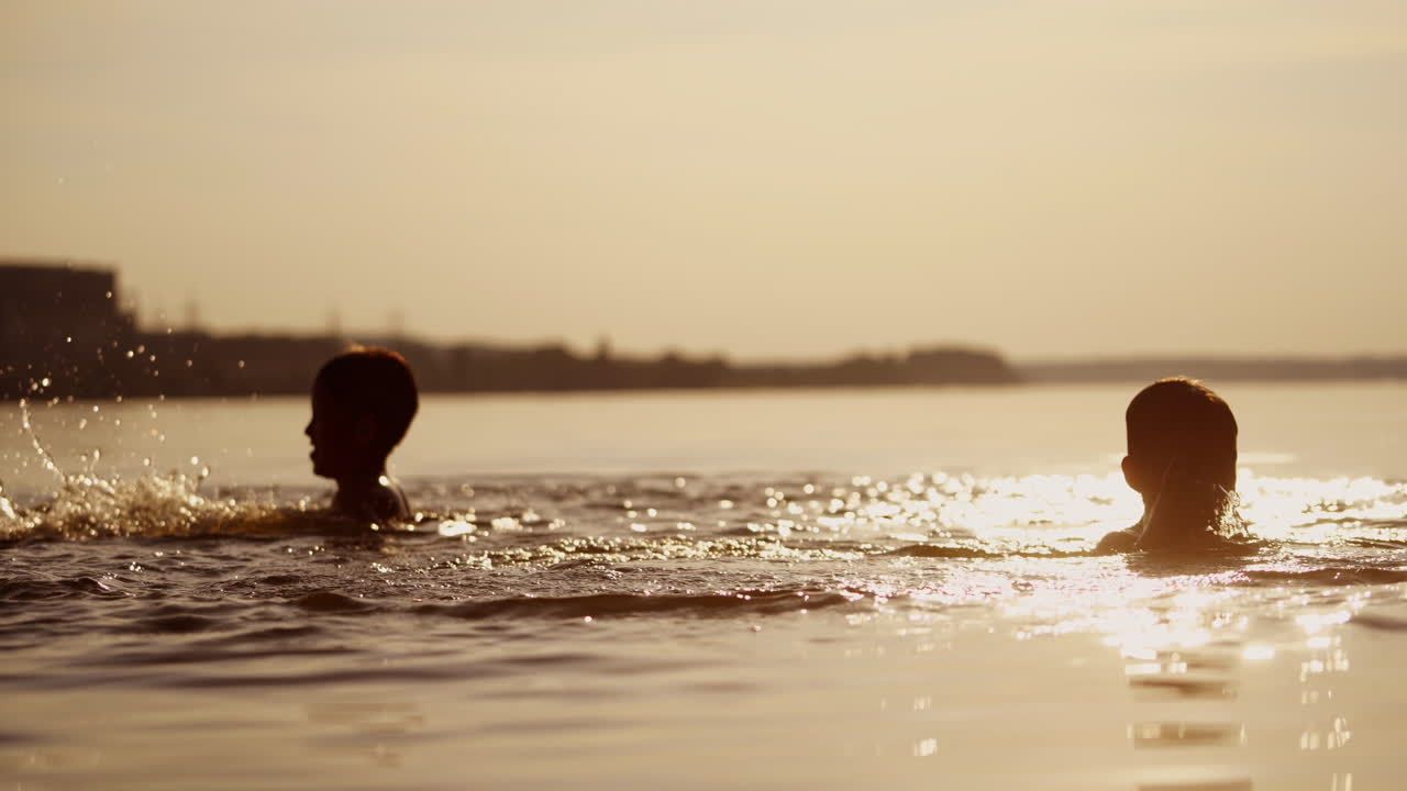 Little kids are playing in water and making splash; Children are swimming in lake or river and having fun with drops; Two brothers rollick at sunset. Monochrome video. Side view.