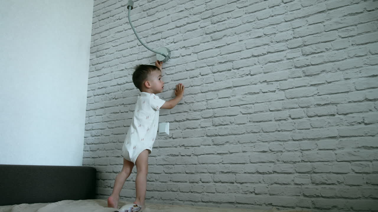 Little toddler boy in a romper runs up to a light switch. Kid turns on the lamp smiling and jumping cheerfully.