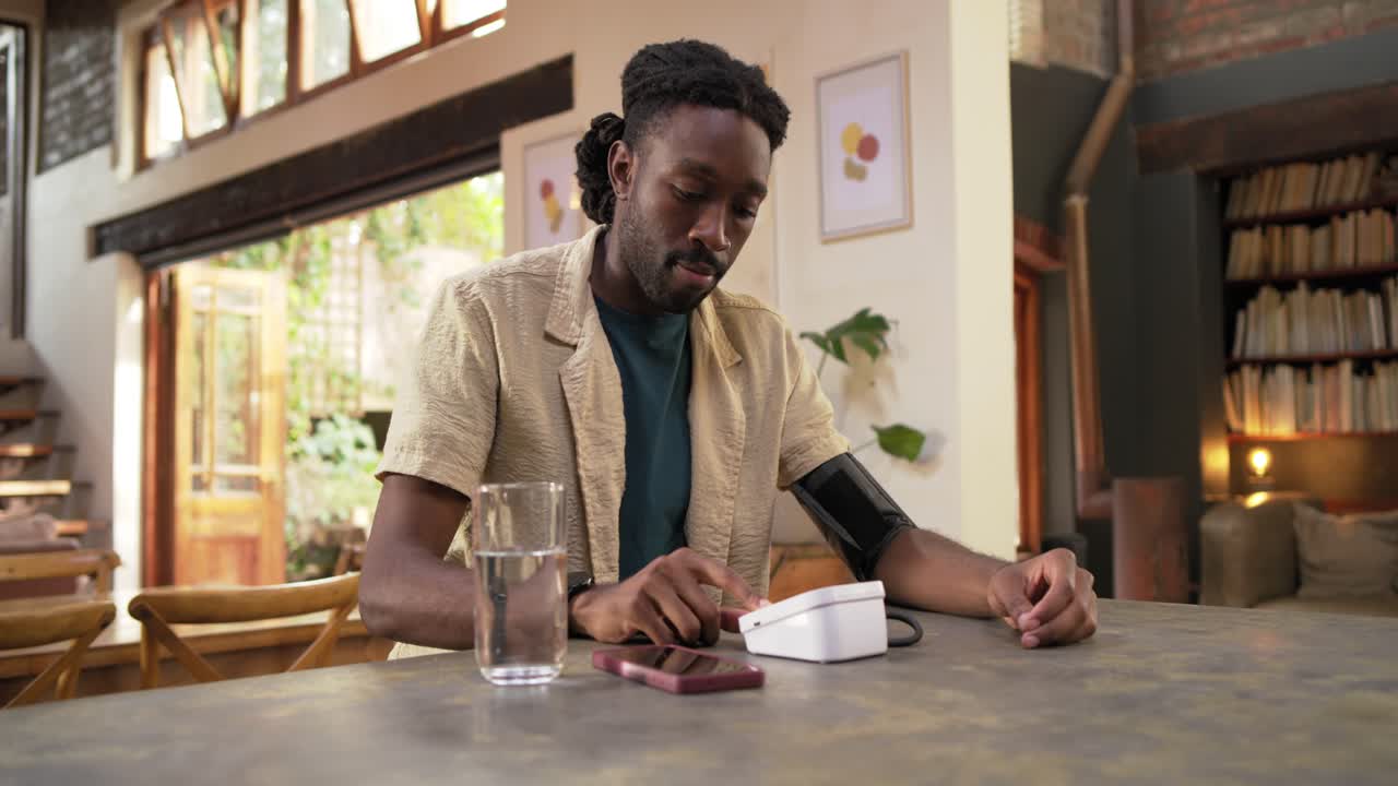 Uomo africano adulto seduto a un tavolo da pranzo in legno che allunga la mano verso il bracciale, iniziando a misurare la pressione sanguigna.