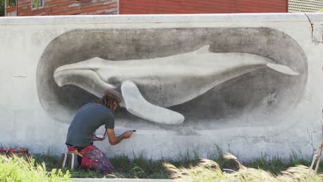 video of caucasian male artist with dreadlocks kneeling painting whale mural on wall