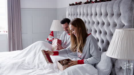 woman opening gift of necklace in bed at home as couple exchange presents on christmas day