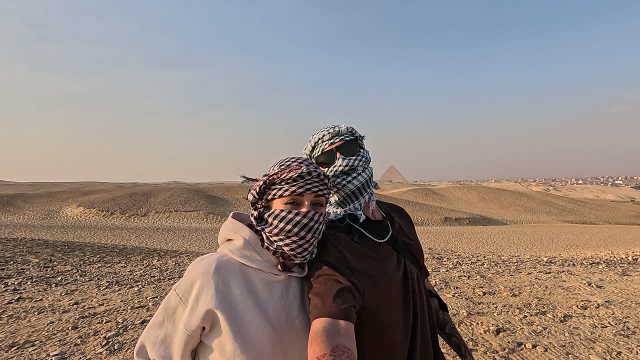 Par iført keffiyeh-tørklæder tager selfie i den egyptiske ørken med pyramider.