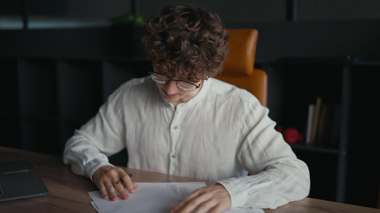 셔츠를 입고 안경을 쓴 곱슬머리를 가진 자신감 있는 젊은 남자가 사무실의 테이블에서 필요한 서류에 서명합니다.