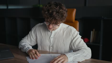 Confident-young-guy-with-curly-hair-wearing-glasses-in-a-white-shirt-signs-the-necessary-papers-at-the-table-in-the-office