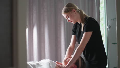 massage therapist in black uniform gently massaging chest of light-skinned female client lying on table under white sheet, serene environment with soft natural light and grey curtain in background