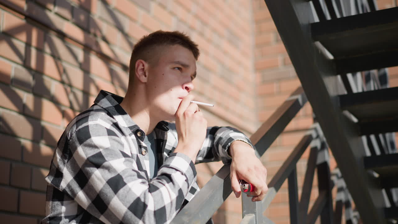 white man leaning on iron railing smoking outdoors holds cigarette in mouth grips lighter and pack in left hand wearing flannel shirt and jeans against bright brick wall under staircase urban vibe