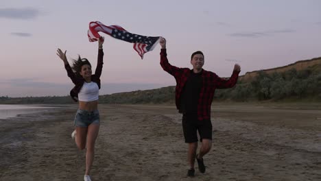 beautiful young two people running on a seaside in the evening. dusk holding above an american flag. happy, smiling, emotions