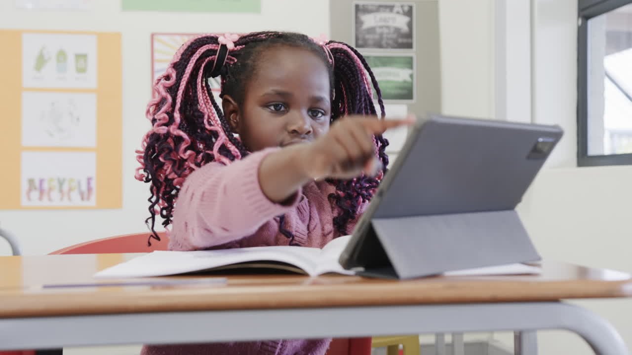 In classroom, African American girl studying with tablet and book, looking focused, at school