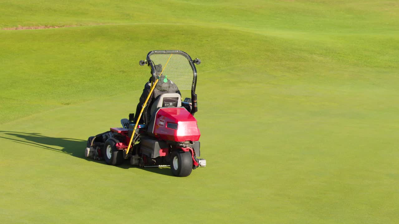 Red riding mower trims green golf course fairway in bright daylight, wide aerial perspective