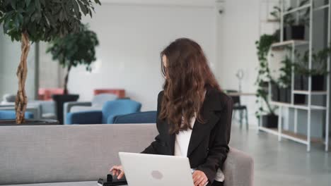 young woman with a laptop, sitting on the couch, is very happy and laughing about the message she received on her smartphone, information about the win