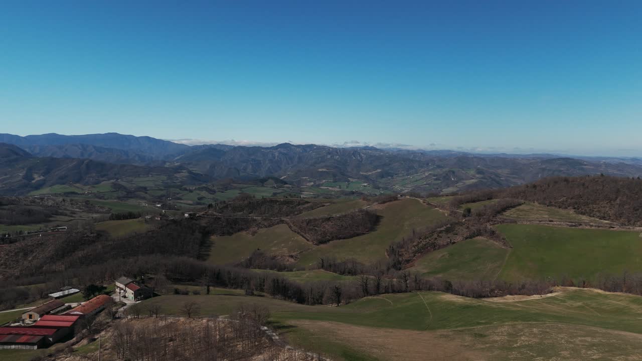 Ampio paesaggio aereo di dolci colline verdi, campi aperti e una catena montuosa distante sotto un cielo azzurro limpido e luminoso