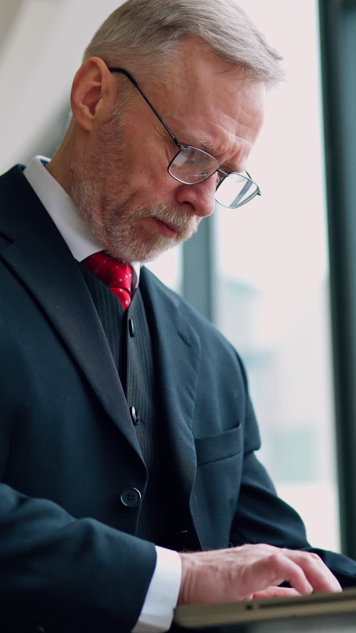 Senior serious businessman standing near window with panoramic city view. Laptop in hands, man thinking while checking e-mails standing near window. Video from the side. Vertical video