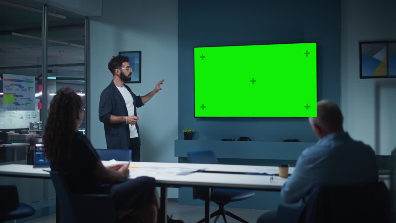 el gerente de operaciones de la compañía realiza una presentación de reunión para un equipo de economistas. hombre adulto usa un televisor ancho con pantalla verde horizontal. la gente trabaja en una oficina de negocios.