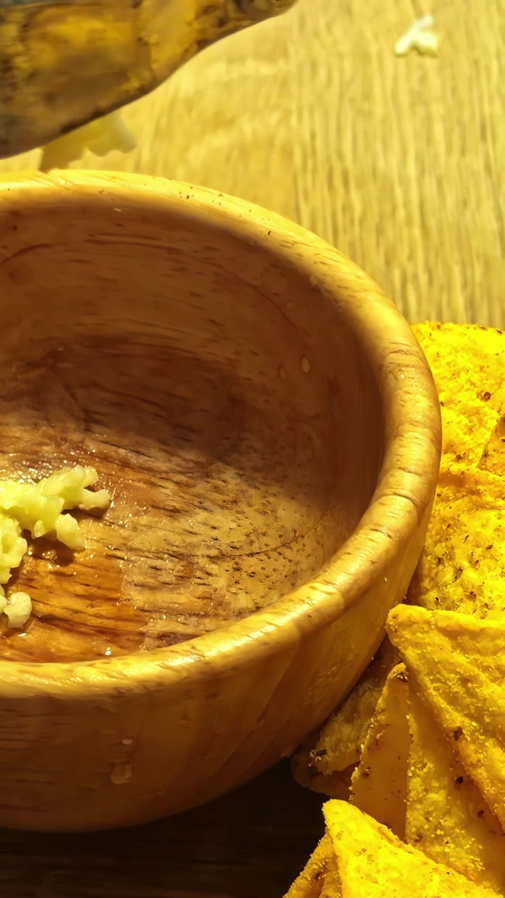Garlic cloves are pressed with a metal garlic press into a wooden bowl placed beside crunchy nacho chips on a wooden surface.