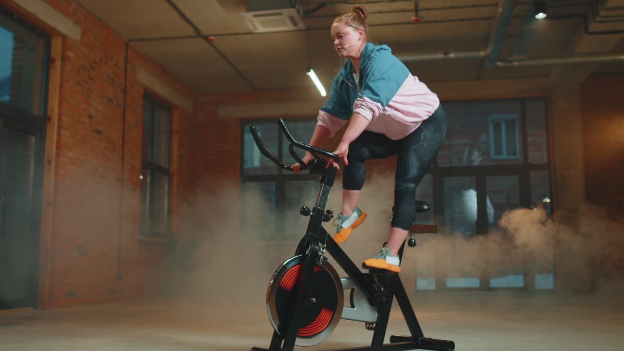 chica atlética realizando ejercicios de entrenamiento de equitación aeróbica en bicicleta estacionaria en un gimnasio con niebla