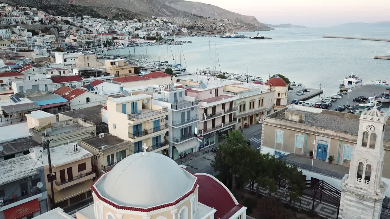 Greece,Kalimnos,Aerial footage of the city center, clock tower and the church behind the iconic municipality building, footage passing these buildings and move towards to the harbor view of the boats.