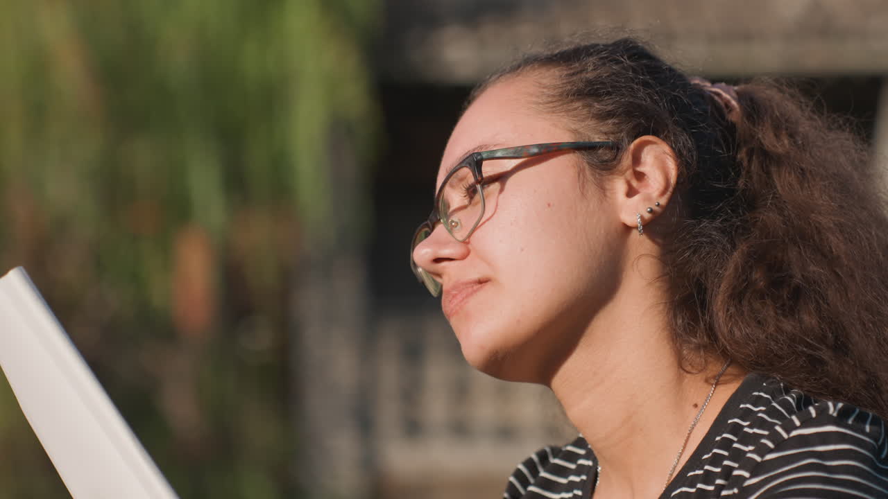 Donna multirazziale che scrive un diario all'aperto all'ora d'oro, tiene vicino il taccuino, camicia a righe, occhiali, sfondo di un cortile urbano, espressioni pensierose, la penna si ferma, luce soffusa posteriore, ritratto intimo