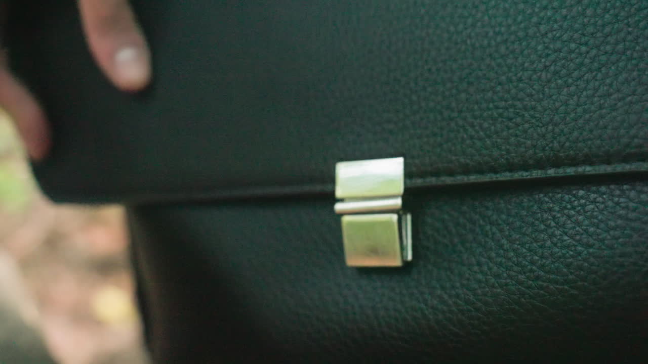 Close-up view of human hand locking and unlocking black leather bag with shiny silver clasp, showing texture of material and movement of fingers, capturing secure action in natural light