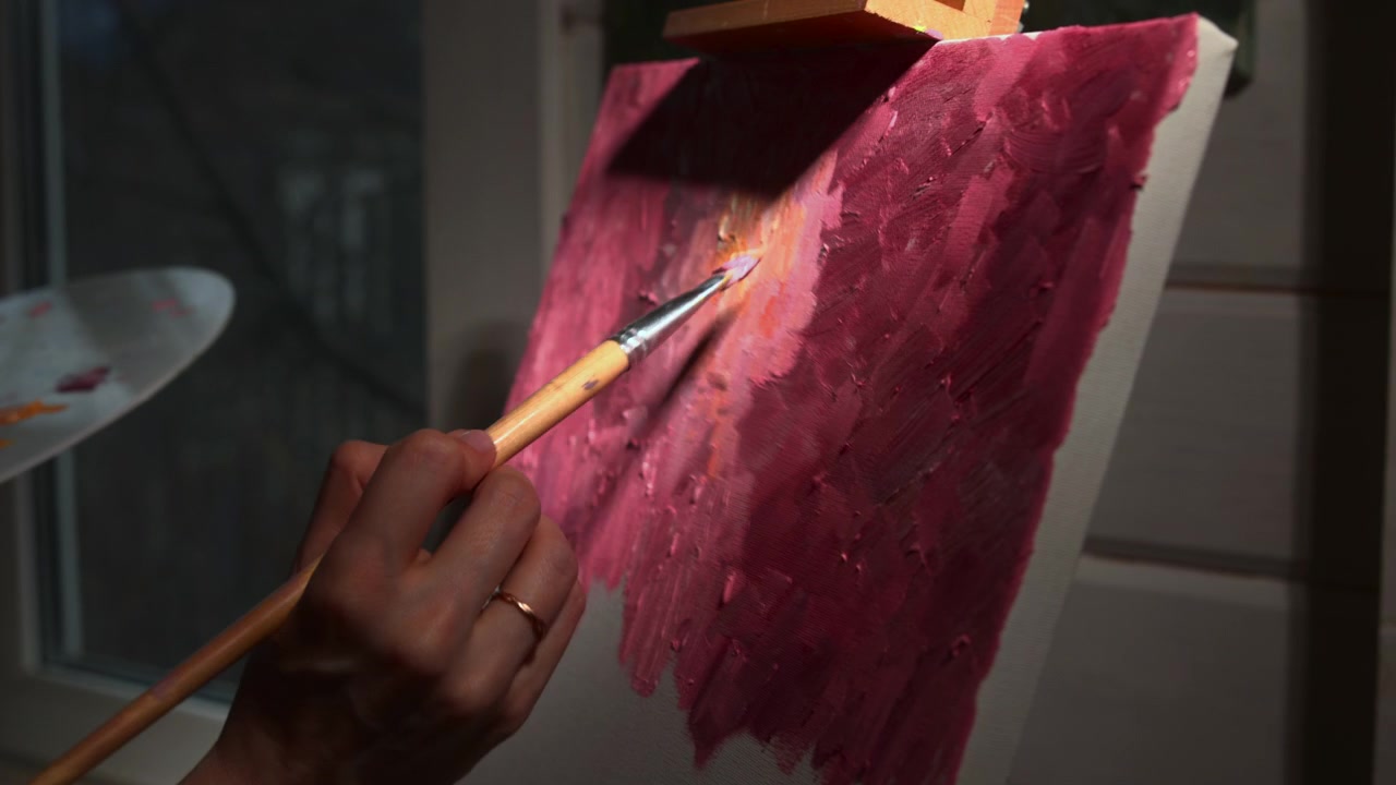 Primo piano di una mano che usa un pennello per applicare pittura rosa su una tela texturizzata all'interno, con una tavolozza visibile nelle vicinanze e una luce calda e soffusa che crea un'atmosfera calma.